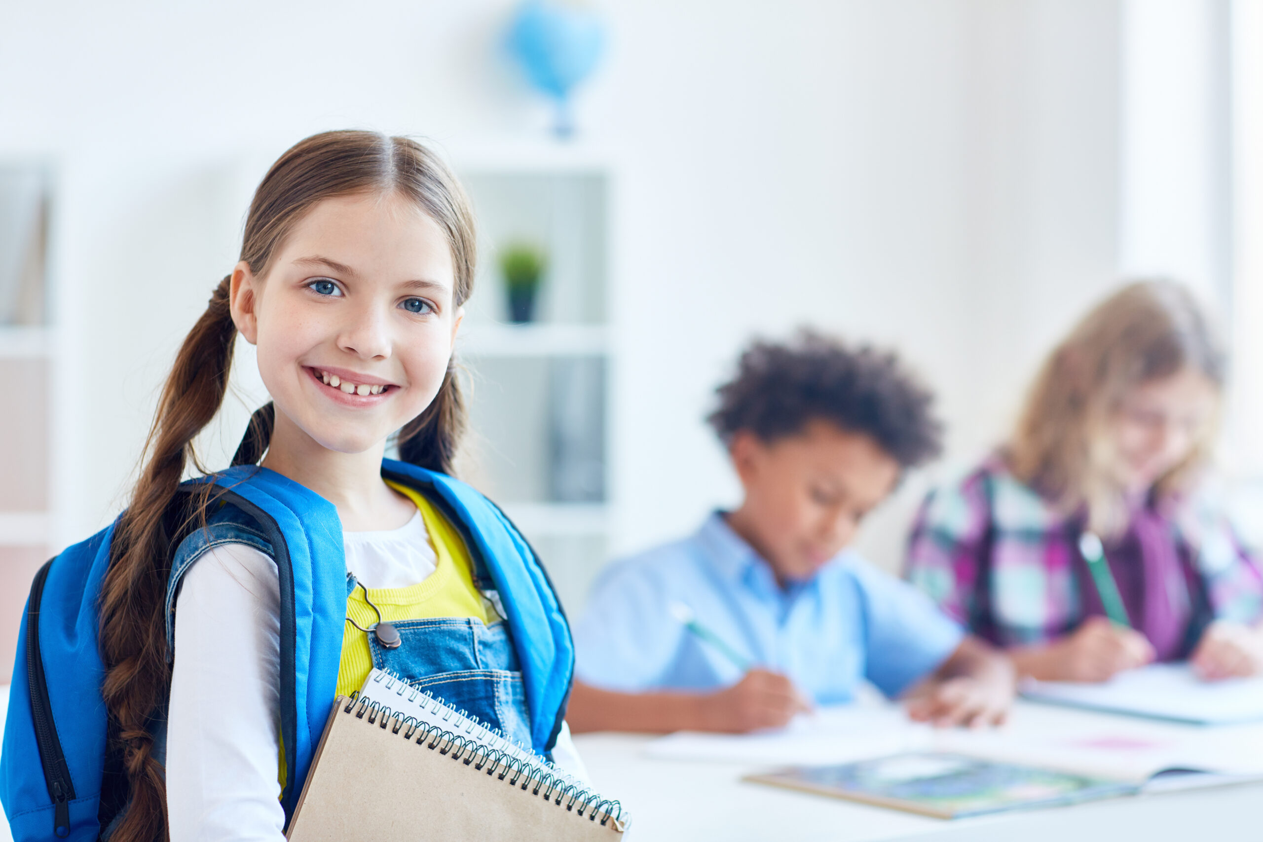 Smiling little scholar with rucksack and notepads on background of schoolboys